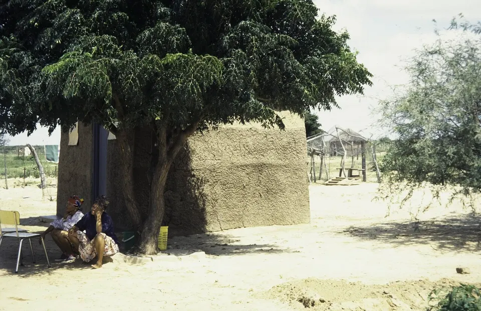 ASC Leiden - van Achterberg Collection - 5 - 065 - Two women sit in the shade of a tree in front of a square house - Groote Laagte, Botswana, 9-15 March 1997 — Ghanzi District, Botswana
